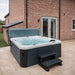 H2O Azure 5-Person Hot Tub installed against a brick house exterior with cover lifter raised and entry steps, dark grey woodgrain cabinet