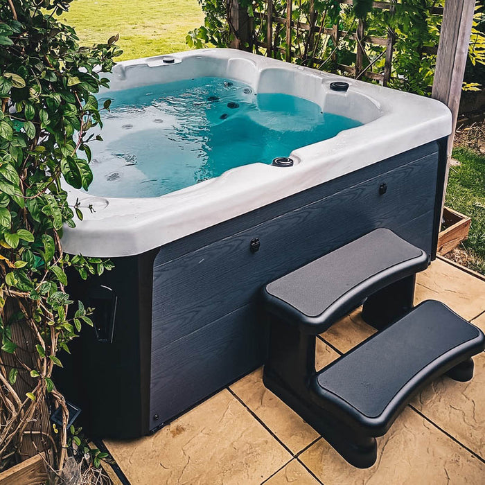 H2O Azure 5-Person Hot Tub on a garden patio with raised wooden border surround, teal blue water, dark grey cabinet and entry steps