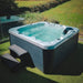 H2O Azure 5-Person Hot Tub on grass in a rural setting with glass screen surround, teal blue water, dark grey cabinet and entry steps