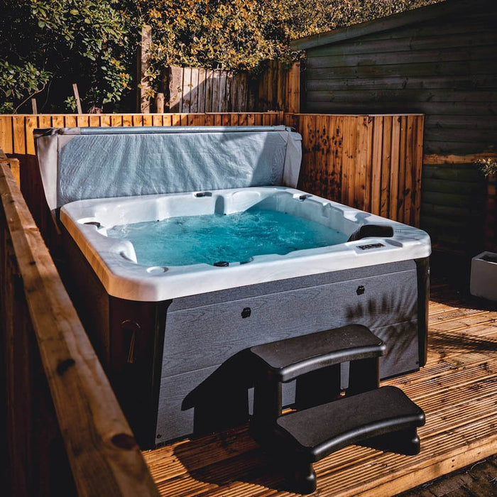 H2O Azure 5-Person Hot Tub on wooden decking at night with cover lifter raised, dark grey woodgrain cabinet, teal water and entry steps