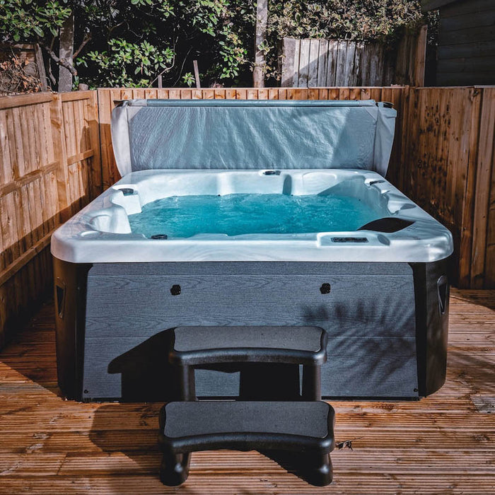 H2O Azure 5-Person Hot Tub on wooden decking at night with cover lifter raised, teal blue water illuminated, dark grey cabinet and entry step.