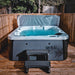 H2O Azure 5-Person Hot Tub on wooden decking at night with cover lifter raised, teal blue water illuminated, dark grey cabinet and entry step.