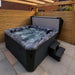H2O Coral 5-Person Hot Tub under a wooden pergola with fountain jets running, cover lifter in use, dark cabinet and steps