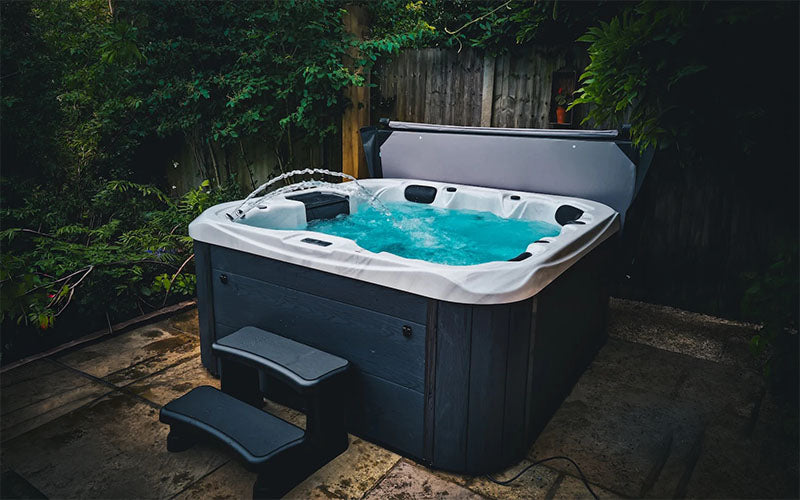 H2O Retreat Plus Hot Tub in grey cabinet installed in a lush garden setting with waterfall feature and black steps.