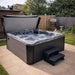 H2O Vortex 6-Person Hot Tub with grey shell and charcoal cabinet, cover open and jets running on a garden patio, shown with entry steps and cover lifter