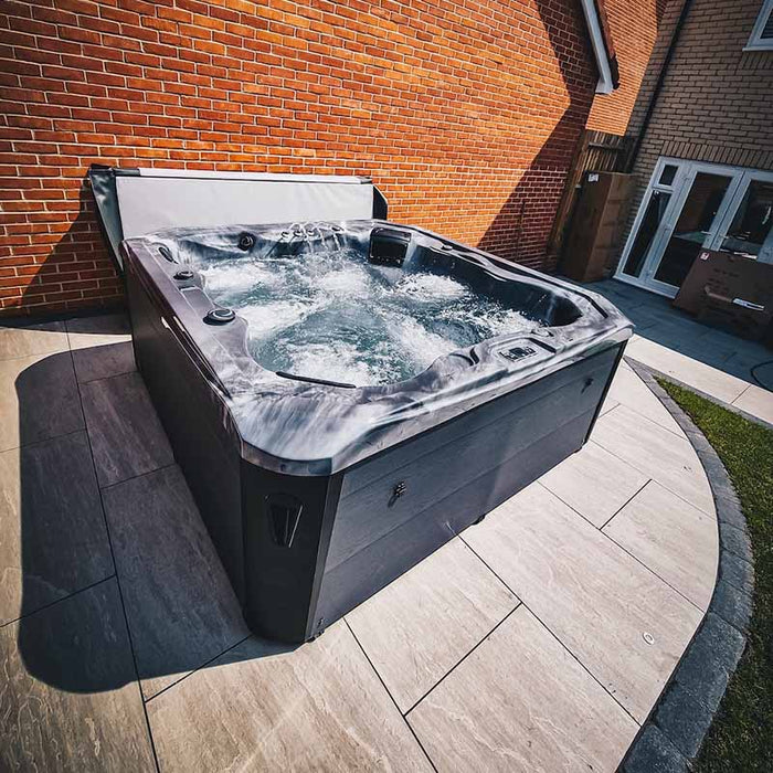 H2O Vortex 6-Person Hot Tub with grey marbled shell and charcoal cabinet, cover open and jets active, installed on an angular stone patio beside a brick house