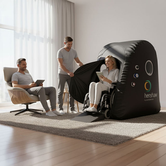 Smiling woman in a wheelchair entering the Henshaw Easy Access Hyperbaric Oxygen Chamber, assisted by family in a relaxed home setting.
