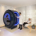 Woman seated inside the Henshaw Monozen Hyperbaric Chamber while a clinician monitors settings, highlighting its clinical use and high-end therapeutic setup.