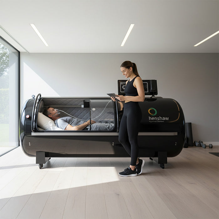 Man receiving oxygen therapy inside the Henshaw Pro Hyperbaric Chamber while a female trainer monitors from outside in a contemporary fitness studio.