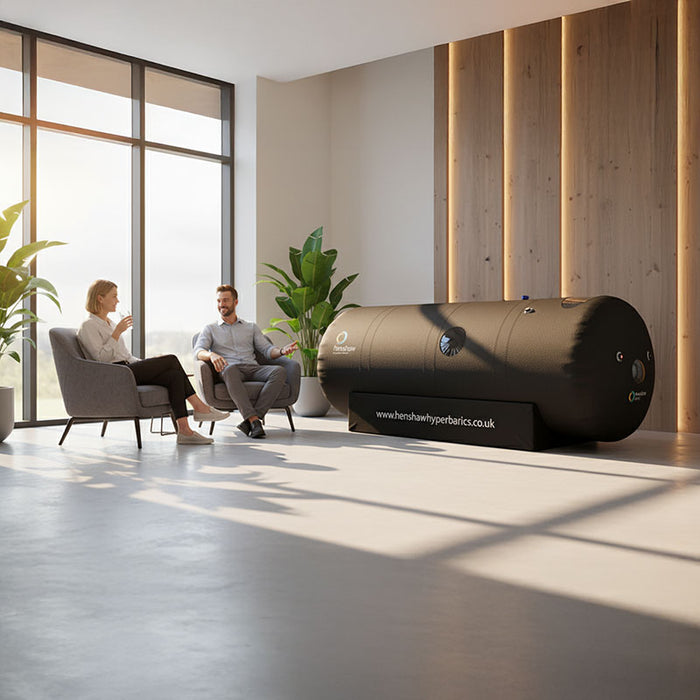 Woman comfortably lying inside the Henshaw Space Hyperbaric Oxygen Chamber while another person supervises, promoting relaxation and recovery.
