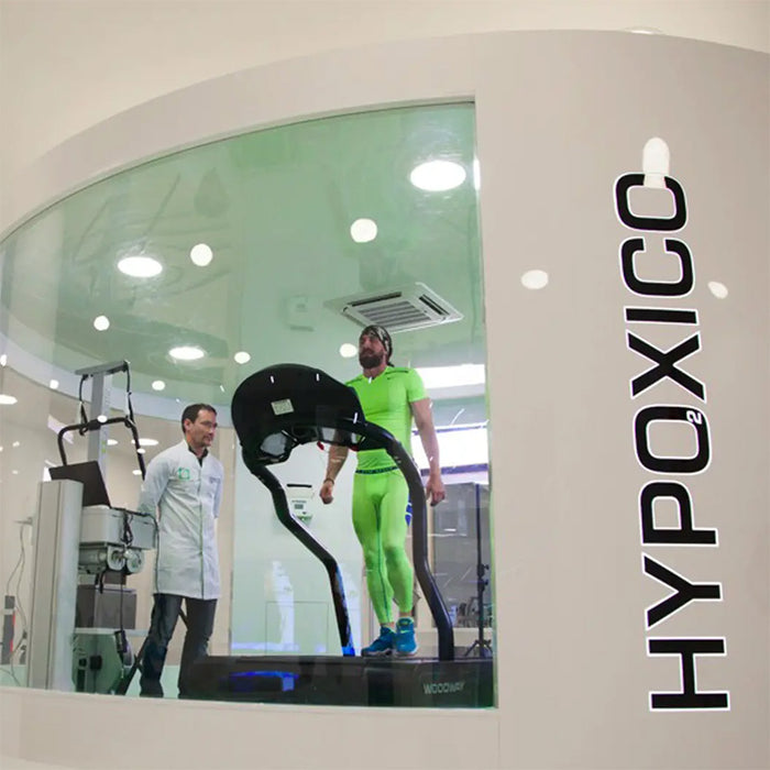 Hypoxico Full Climate Hypoxic Chamber with athlete in green kit on Woodway treadmill inside glass-walled enclosure, supervised by technician in white lab coat.