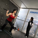 Hypoxico Hypoxic Altitude Chamber altitude room with two athletes on Life Fitness elliptical cross-trainers, wall-mounted display showing 2954m, alpine mountain murals in background.