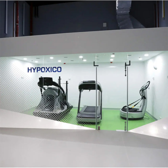 Hypoxico Hypoxic Altitude Chamber compact single-user acrylic pod with aluminium frame containing a Tour de France branded stationary bike, gym strength equipment in background.