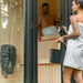 Woman entering the Jonas Classic 4-person sauna holding a wooden sauna bucket, with man seated inside on the upper bench.