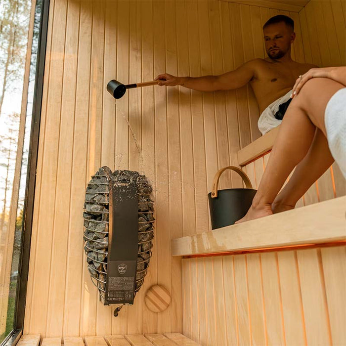Man pouring water onto HUUM heater while relaxing in the Jonas Classic sauna with a woman seated beside him on upper bench.