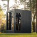 Side profile of the Jonas Classic outdoor sauna set amongst trees, highlighting tall reflective glass panels and clean architectural lines.