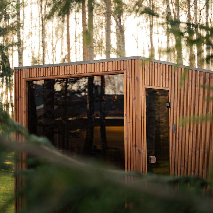 Jonas Grand 8-person garden sauna nestled in a forest landscape, showcasing its sleek design with natural timber finish and large glass front.