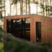 Jonas Grand 8-person garden sauna nestled in a forest landscape, showcasing its sleek design with natural timber finish and large glass front.