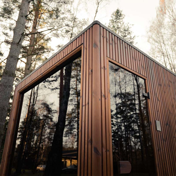 Close-up view of the Jonas Grand 8-person outdoor sauna showing sleek corner detailing and large reflective glass panels set in natural wood surroundings.