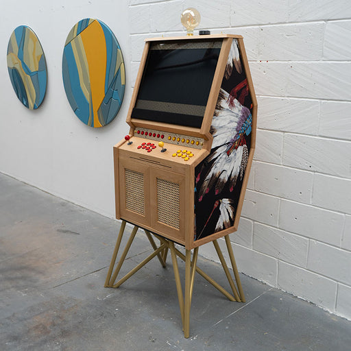 Maison Roshi Senpai V3 arcade cabinet with retro-inspired controls, wooden frame, feather artwork on side panels, and golden tripod legs in modern gallery space.
