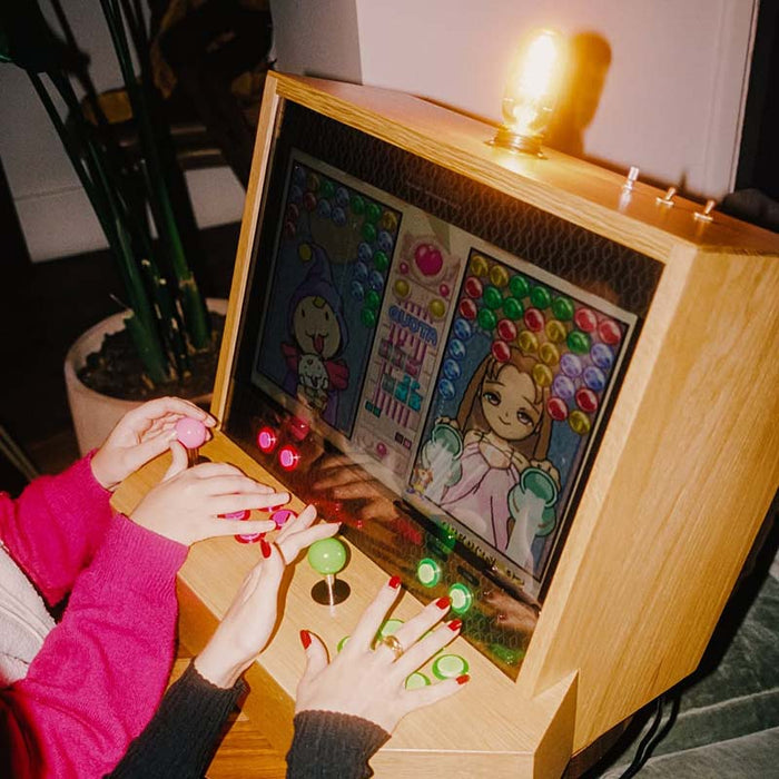 Maison Roshi Sensei V1 arcade cabinet with two players using green and pink retro-style joysticks and colourful buttons on a wooden console, shown in a cosy home setting.