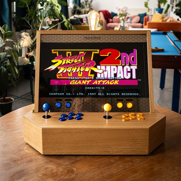 Maison Roshi Sensei V1 arcade console with Street Fighter III: 2nd Impact on screen, blue and orange controls, and a rustic wooden cabinet in a plant-filled room.
