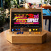 Maison Roshi Sensei V1 arcade console with Street Fighter III: 2nd Impact on screen, blue and orange controls, and a rustic wooden cabinet in a plant-filled room.