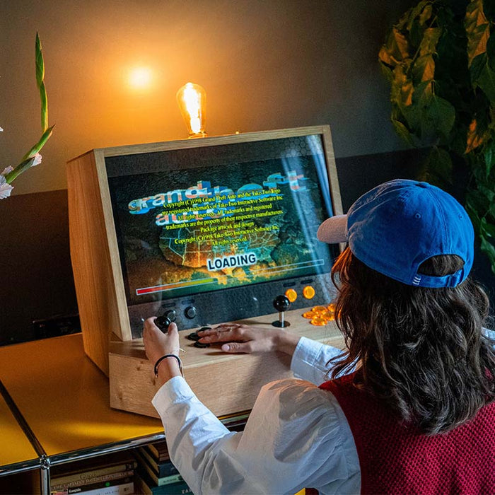 Maison Roshi Sensei V2 arcade cabinet showing Grand Theft Auto loading screen, with glowing filament bulb and black joysticks in a warm ambient room.