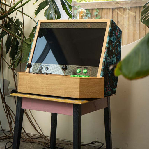 Maison Roshi Sensei V2 retro arcade console with green and black joysticks, styled with decorative fabric side panels, standing in a leafy home corner.