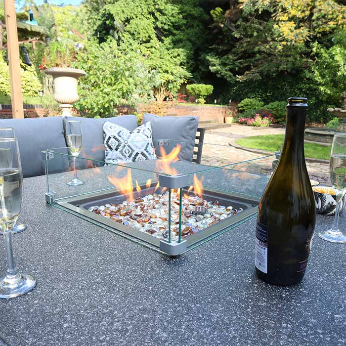 The Mayfair Corner Lounge Set's fire pit table glowing in an outdoor garden setting, with comfortable seating and a bottle of wine, creating a perfect atmosphere for relaxing in nature.