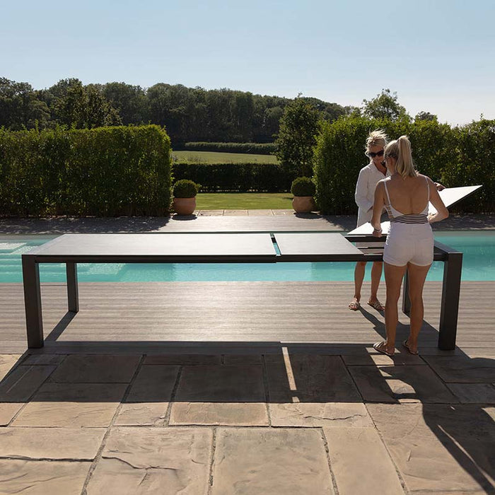 Two women setting up the Ambition 10 seater extending dining table in oatmeal, positioned by the pool for outdoor entertaining.