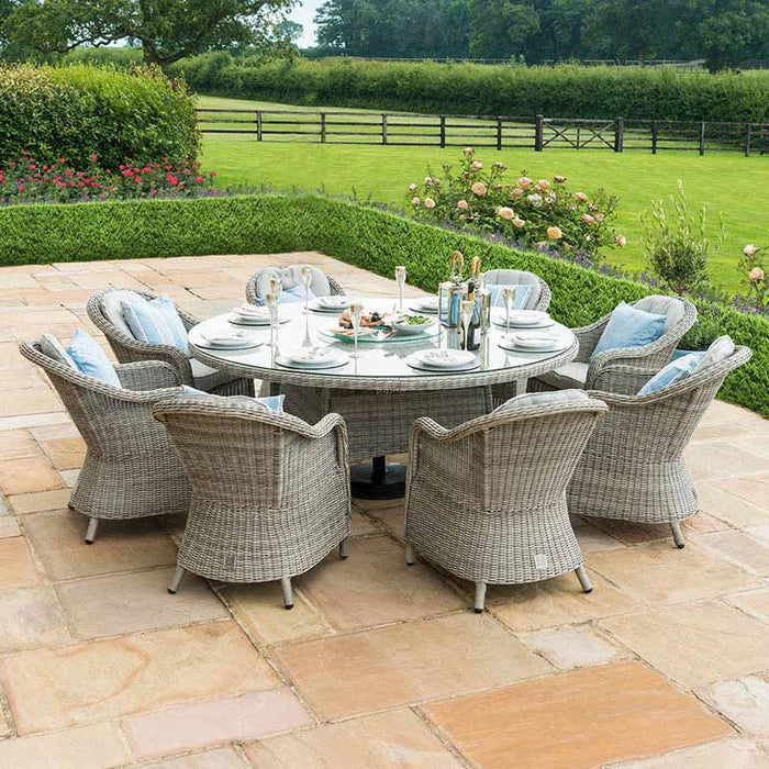 Maze Oxford Heritage 8 Seater Round Dining Set displayed on a patio, complete with grey rattan chairs and a glass-top table, set amidst a scenic garden.