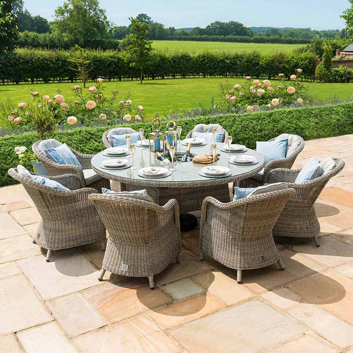 The Maze Oxford Heritage 8 Seater Round Dining Set, arranged on a patio, featuring grey rattan chairs with blue pillows and a glass-topped table, set against a lush garden backdrop.