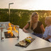 Woman and child chatting by the Pulse 3 Seater Sofa Dining Set with Fire Pit, flames glowing and snacks served on the table.