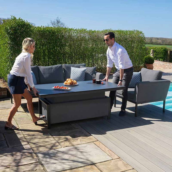 Two people adjusting the height of the Maze Pulse 3 Seater Sofa Dining Set with Rising Table in flanelle. The spacious outdoor set, located poolside, offers flexibility for both dining and relaxing, with the table smoothly transitioning between coffee table and dining height.