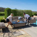 A couple adjusts the rising table’s height, showcasing the practical functionality of the Maze Pulse 3 Seater Sofa Dining Set in Taupe for dining or lounging outdoors.