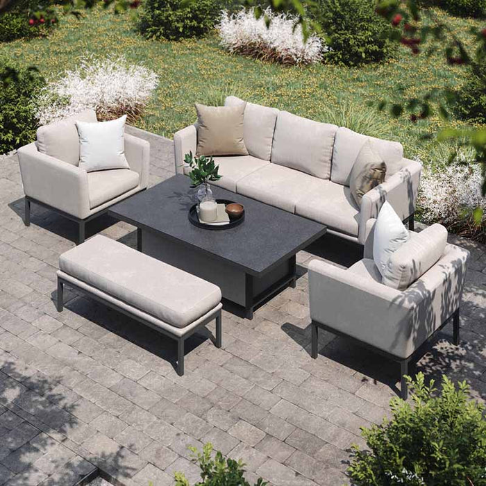 A stylish outdoor setup featuring the Maze Pulse 3 Seater Sofa Dining Set in Oatmeal, complete with a rectangular rising table, armchairs, and a bench, arranged on a stone patio amidst lush greenery.