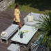 A woman walking beside the Pulse Rectangular Corner Dining Set with Rising Table in Lead Chine, showcasing the elegant and inviting outdoor setup in a serene garden setting.