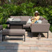 Woman relaxing on the Maze Pulse Square Corner Dining Set with Fire Pit in taupe, set in a lush outdoor garden with modern cushions, creating a serene and stylish lounging experience.