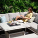 Outdoor Pulse Square Corner Dining Set with Rising Table in a garden setting. Woman relaxing on a grey cushioned corner sofa, featuring a square adjustable table for versatile dining and lounging.