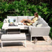 Woman relaxing on the Pulse Square Corner Dining Set with Rising Table, surrounded by cushions and greenery in lead chine.