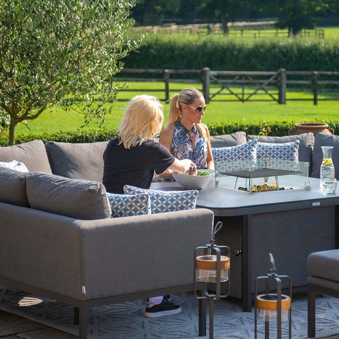 Two women enjoying the Pulse U Shape Corner Dining Set with Fire Pit in a luxurious garden setting.