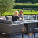 Two women enjoying the Pulse U Shape Corner Dining Set with Fire Pit in a luxurious garden setting.