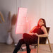 Woman enjoying a relaxing red light therapy session with the Nuvibody LITE 1500 panel mounted on an adjustable stand, ideal for skin rejuvenation and half-body treatment at home.