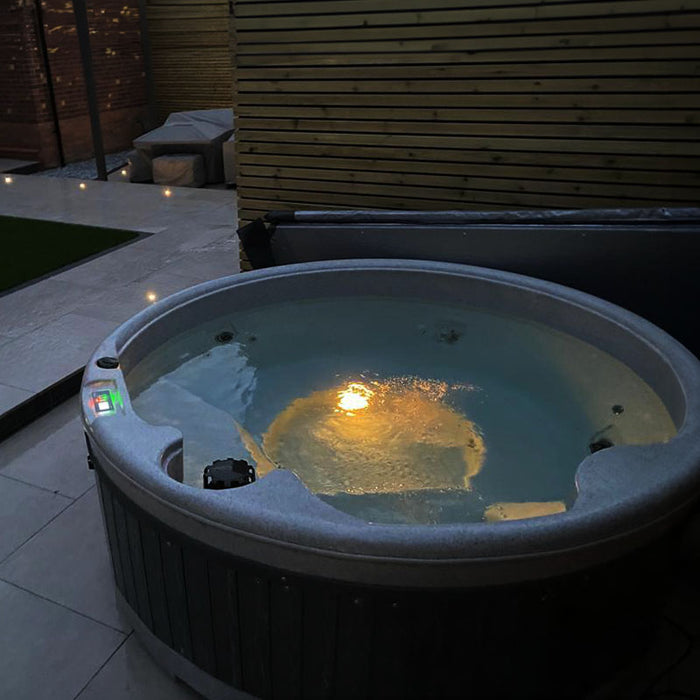 Grey hot tub with yellow lighting in an outdoor patio at night.