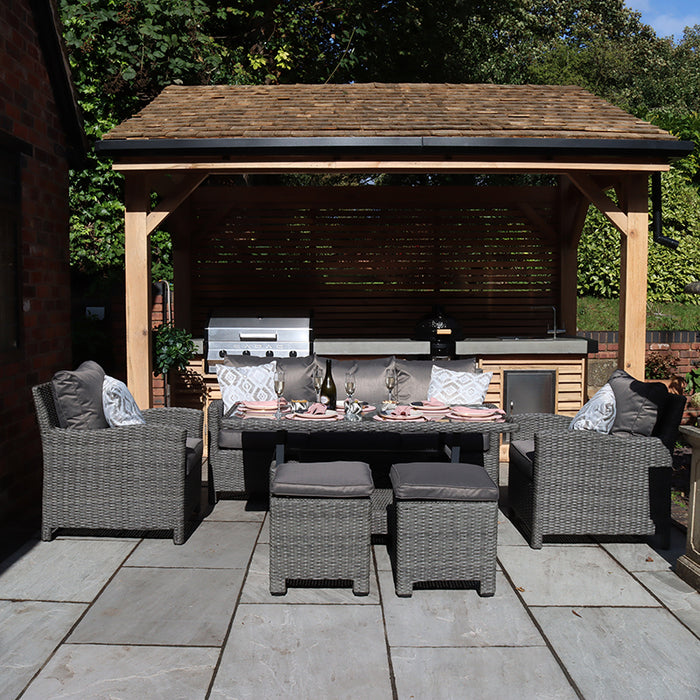 A welcoming outdoor scene with the Paris 7 Seater Deluxe Sofa Dining Set, including a spacious sofa, armchairs, stools, and a glass-top dining table, all arranged under a wooden structure for a perfect garden retreat.