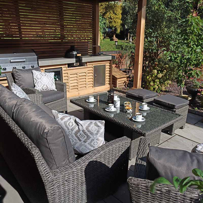 The Paris 7 Seater Deluxe Sofa Dining Set arranged under a wooden gazebo, showcasing a sleek glass dining table set for coffee. The durable grey wicker and plush cushions enhance the garden's modern yet cosy vibe.