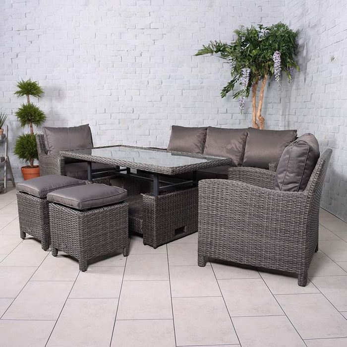 The Paris 7 Seater Deluxe Sofa Dining Set arranged in an indoor corner, featuring a glass-top dining table, cushioned grey wicker armchairs, and stools, creating a stylish and comfortable seating area.