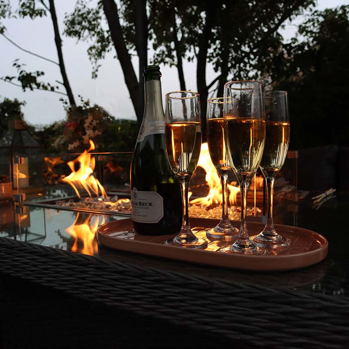 Champagne glasses and a bottle on the Paris Fire Pit Corner Dining & Lounge Set’s rectangular table, illuminated by the fire pit's soft glow.