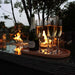 Champagne glasses and a bottle on the Paris Fire Pit Corner Dining & Lounge Set’s rectangular table, illuminated by the fire pit's soft glow.